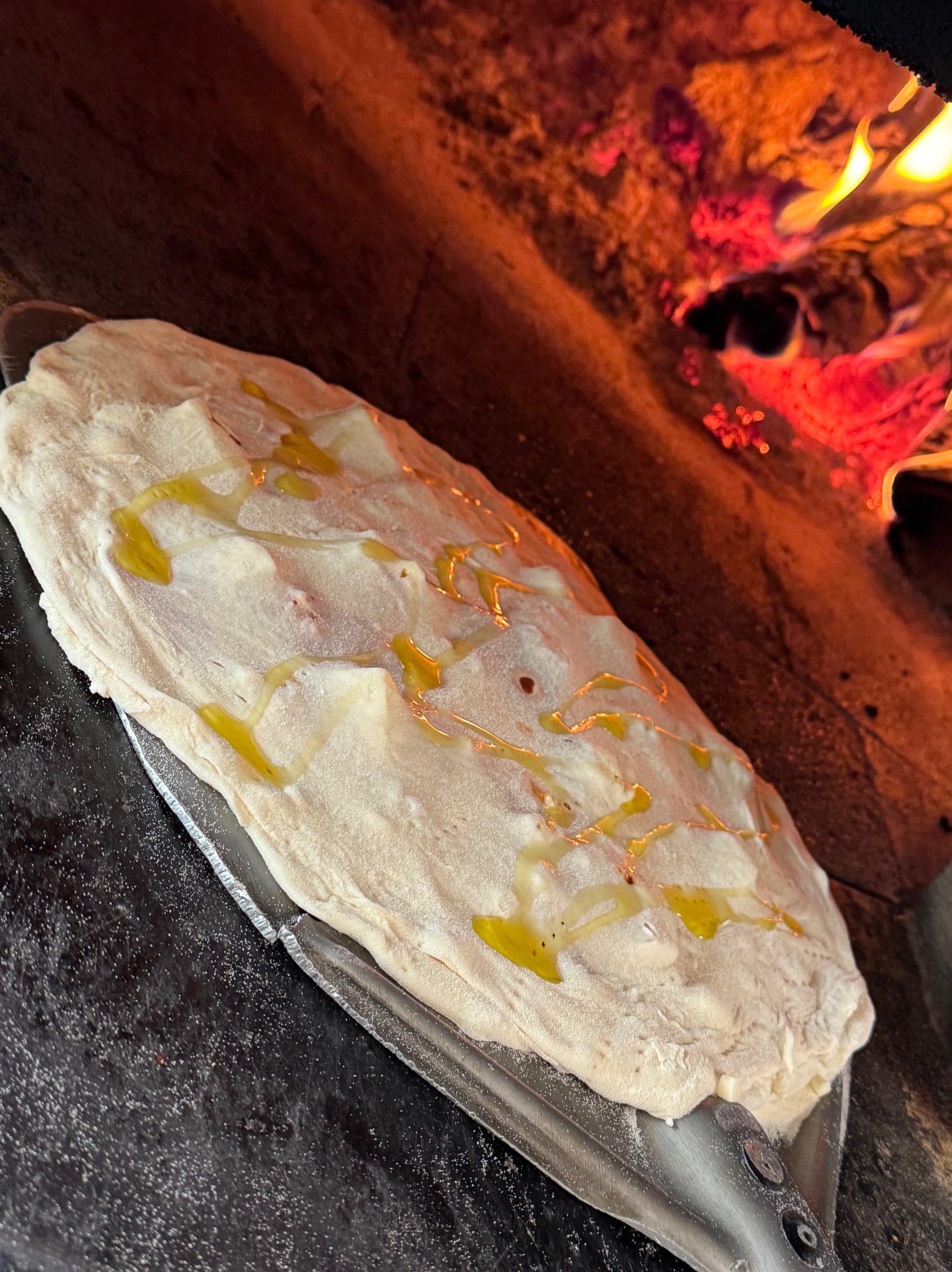 Surowe ciasto na pizzę z oliwą na metalowej łopacie, gotowe do włożenia do pieca opalanego drewnem z żywym ogniem w tle. Przygotowanie pizzy.