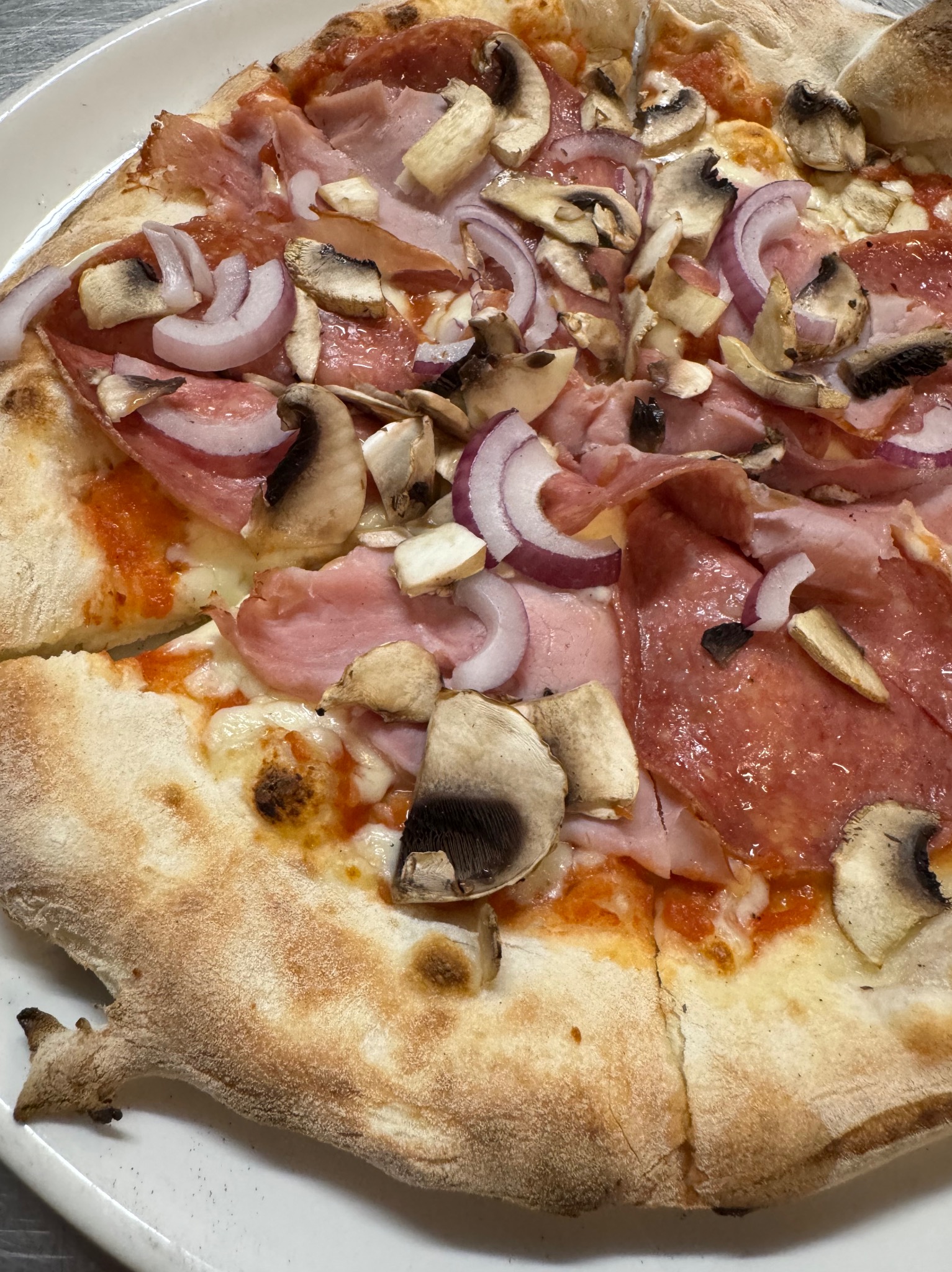 Pizza z szynką, pieczarkami i czerwoną cebulą na talerzu. Widoczne ciasto z pęcherzykami powietrza i apetyczne dodatki. Idealna na kurs kulinarny.