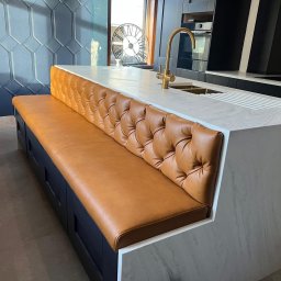 Veluro Jakub Sroka - Wyspa kuchenna z tapicerowanym siedziskiem w kolorze karmelowym, pikowana w romby, na tle ciemnej zabudowy i dekoracyjnej ściany. Złota bateria kuchenna.