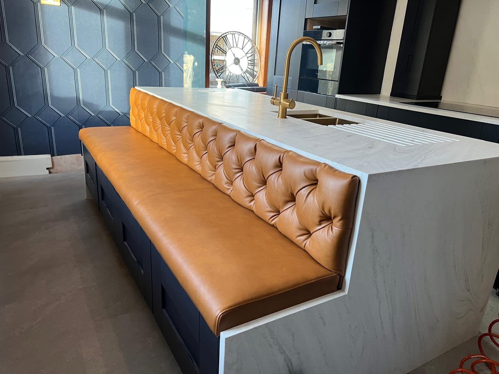 Wyspa kuchenna z tapicerowanym siedziskiem w kolorze karmelowym, pikowana w romby, na tle ciemnej zabudowy i dekoracyjnej ściany. Złota bateria kuchenna.