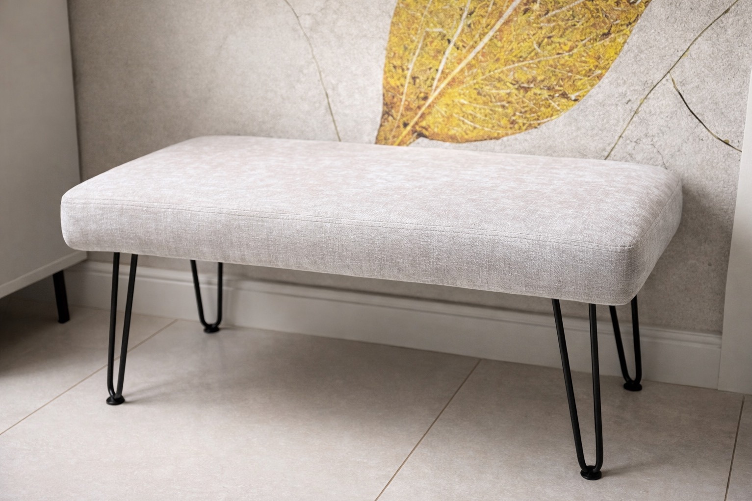 Tapicerowany puf na czarnych, metalowych nóżkach w kształcie litery V, na tle ściany z motywem liścia. Minimalistyczny design, jasna kolorystyka.