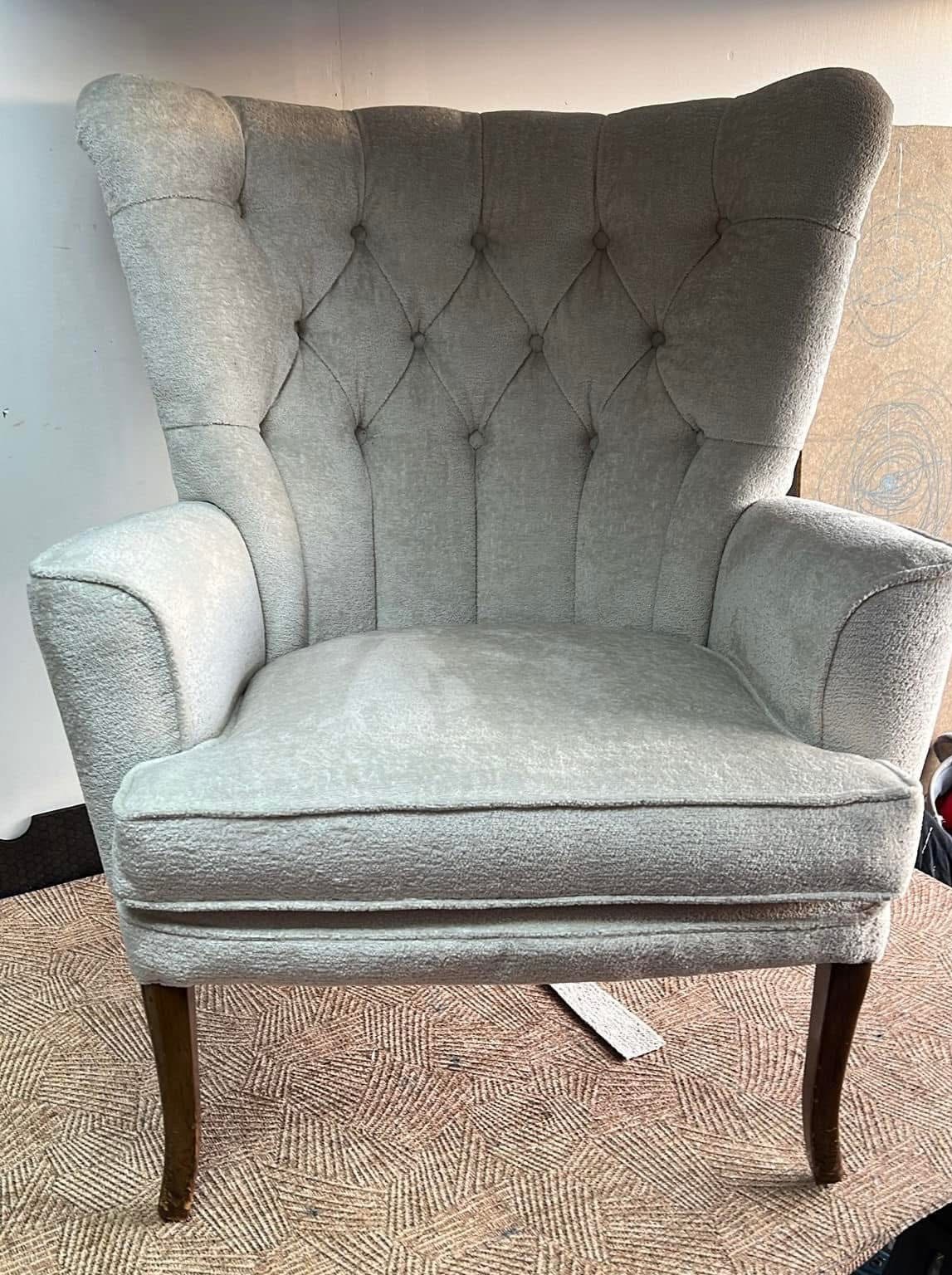 Elegancki, szary fotel typu uszak z pikowanym oparciem, na ciemnych, drewnianych nogach, ustawiony na wzorzystym dywanie. Widoczne detale tapicerki i kształt fotela.