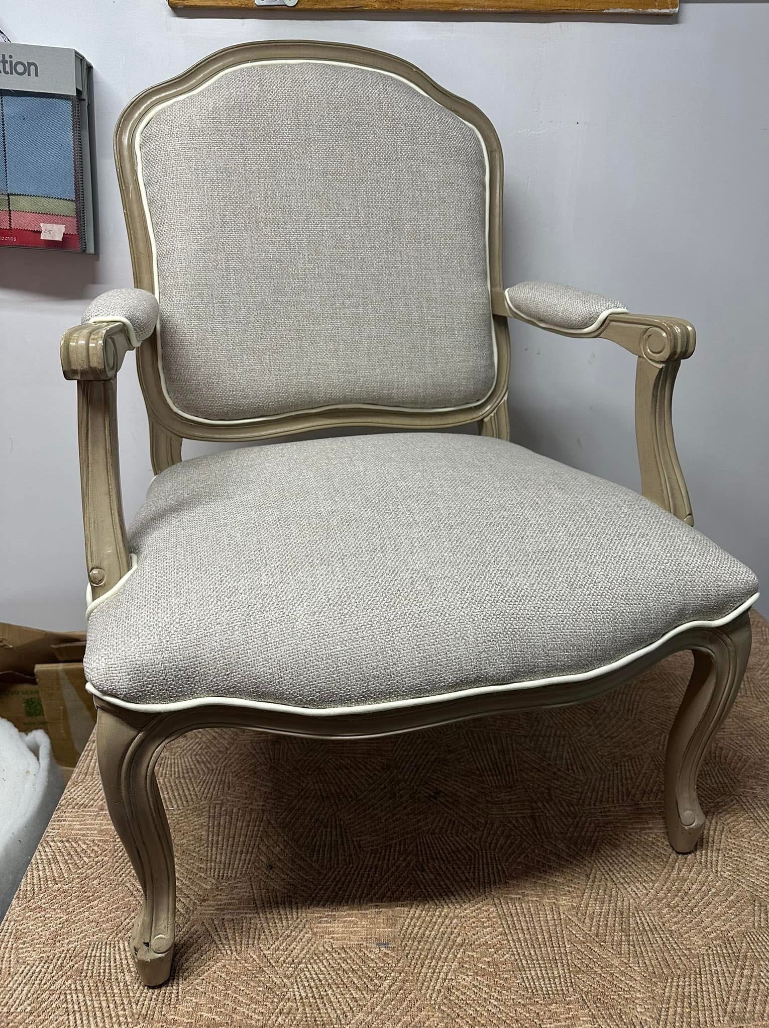 Odnowione krzesło w stylu Ludwika XVI z szarą tapicerką i kremową ramą, wykończone białą lamówką. Widoczny wzornik tkanin w tle.