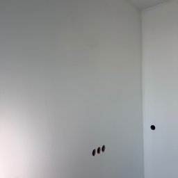 SEBASTIAN PIEROG - Biała ściana w rogu pomieszczenia z trzema otworami na gniazdka elektryczne po lewej i dwoma otworami po prawej stronie. Świeżo pomalowana powierzchnia.