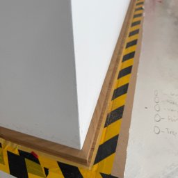 SEBASTIAN PIEROG - Detal wykończenia ściany przy podłodze, zabezpieczony taśmą ostrzegawczą. Widoczne listwy przypodłogowe i notatki na podłodze. Ujęcie z bliska, podkreślające precyzję.