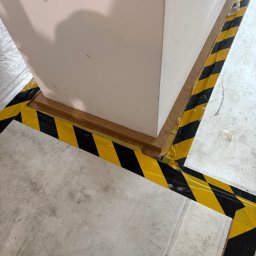 SEBASTIAN PIEROG - Oznakowanie podłogi taśmą ostrzegawczą wokół obudowy kolumny. Fragment podłogi betonowej. Detal wykończenia z drewnianą listwą.