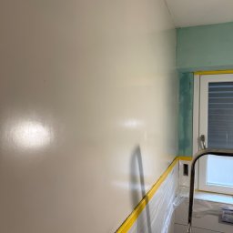 SEBASTIAN PIEROG - Ściana po malowaniu na gładko, zabezpieczona taśmą malarską i folią. Widoczny fragment drabiny malarskiej i okno. Świeża, jasna farba na ścianie.