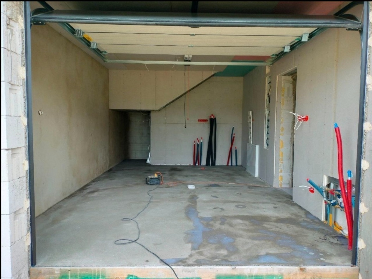 Wnętrze garażu w trakcie adaptacji: surowe ściany, instalacje wodne i elektryczne w ścianach z płyt kartonowo-gipsowych, betonowa posadzka, widoczne narzędzia.