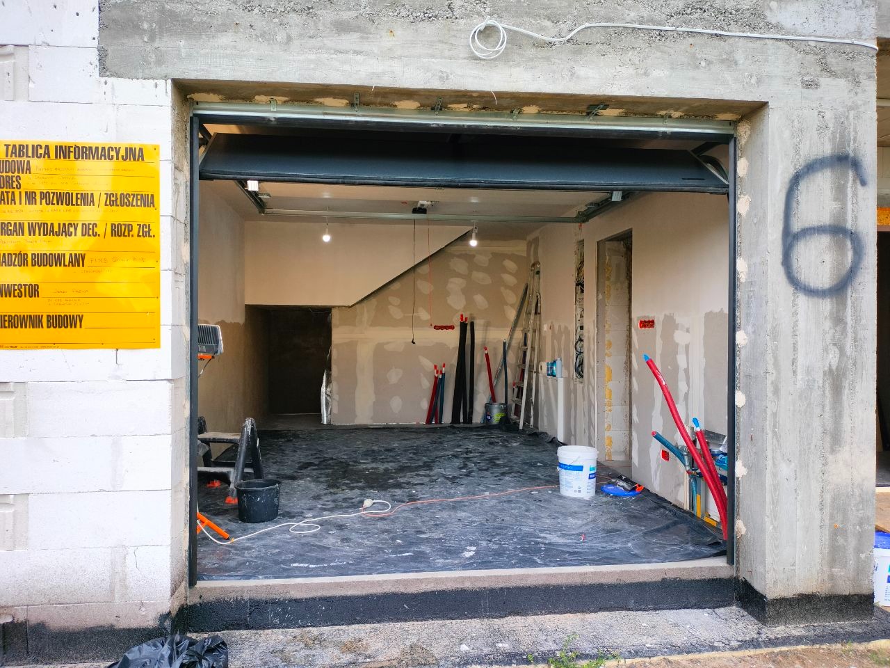 Wnętrze garażu w trakcie prac wykończeniowych: ściany przygotowane do malowania, widoczne instalacje, podłoga zabezpieczona folią. Tablica informacyjna budowy po lewej.