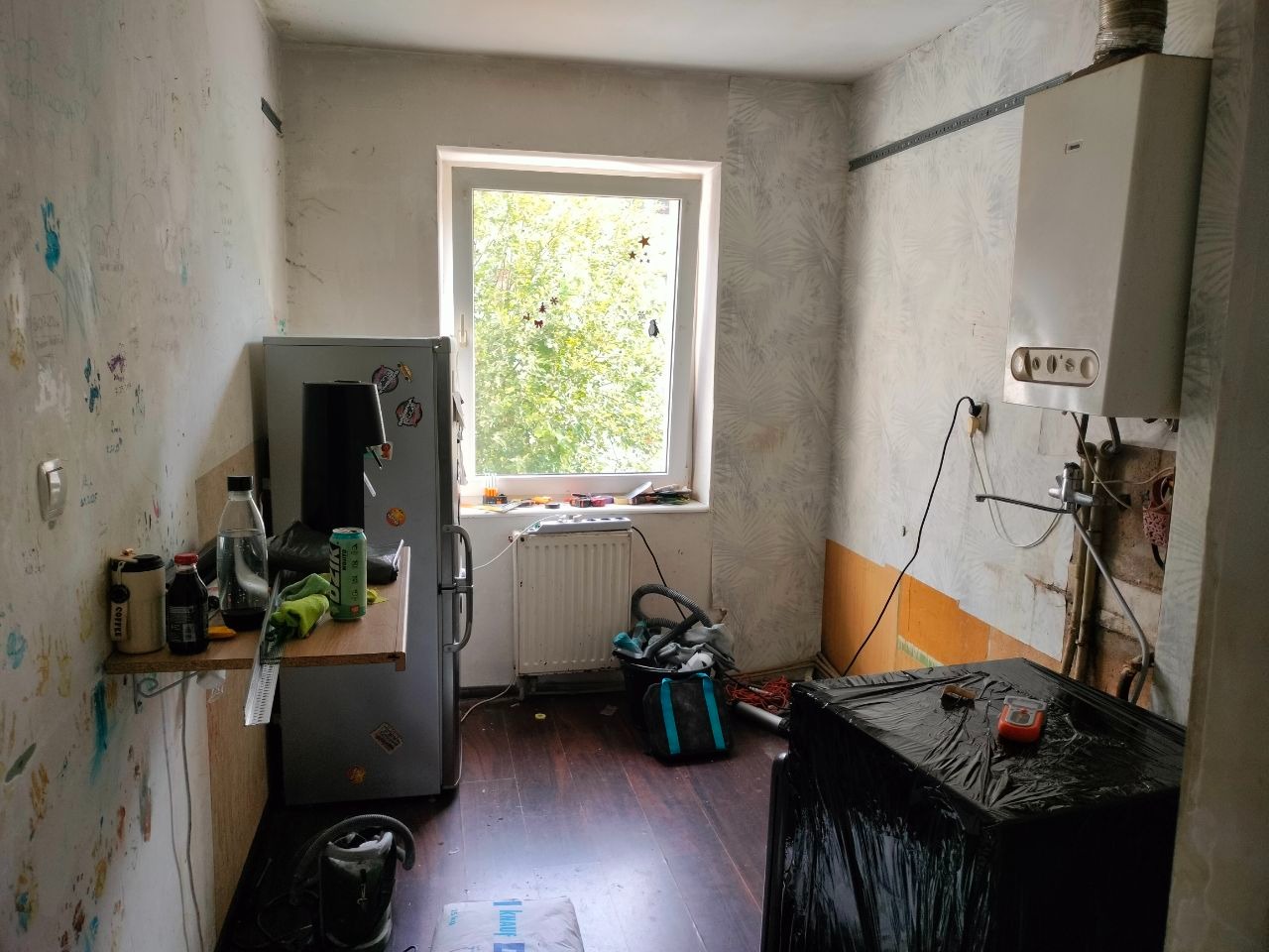 Kuchnia w trakcie remontu: ściany częściowo z tapetą w liście, częściowo z dziecięcymi rysunkami, odsłonięte fragmenty ścian, zakryte meble, okno z widokiem na zieleń.