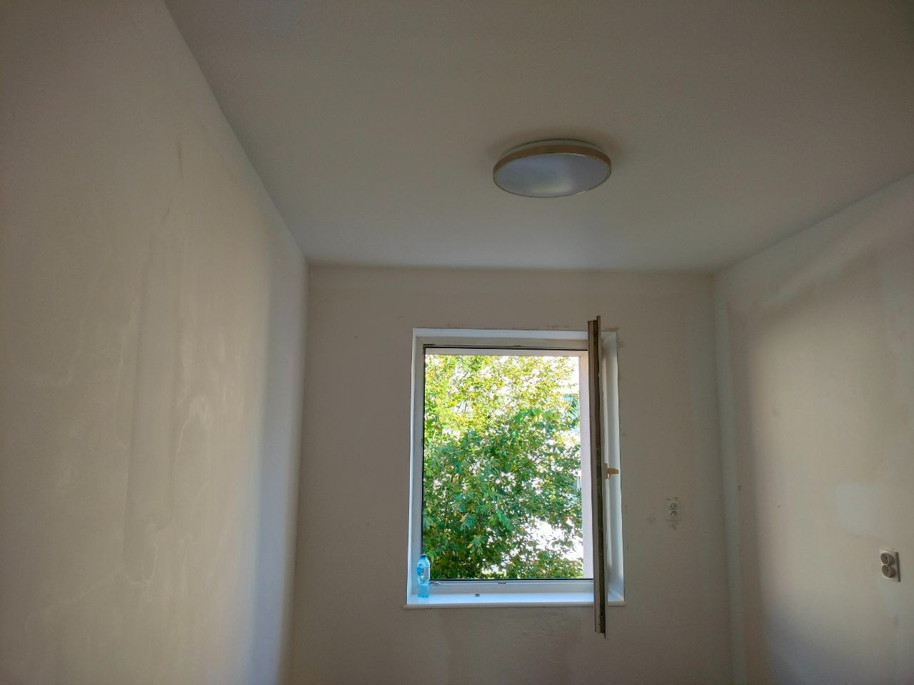 Pokój po szpachlowaniu ścian, przygotowany do malowania. Widoczne ślady po szpachli, okno z widokiem na zieleń i lampa sufitowa. Przygotowanie do malowania ścian.