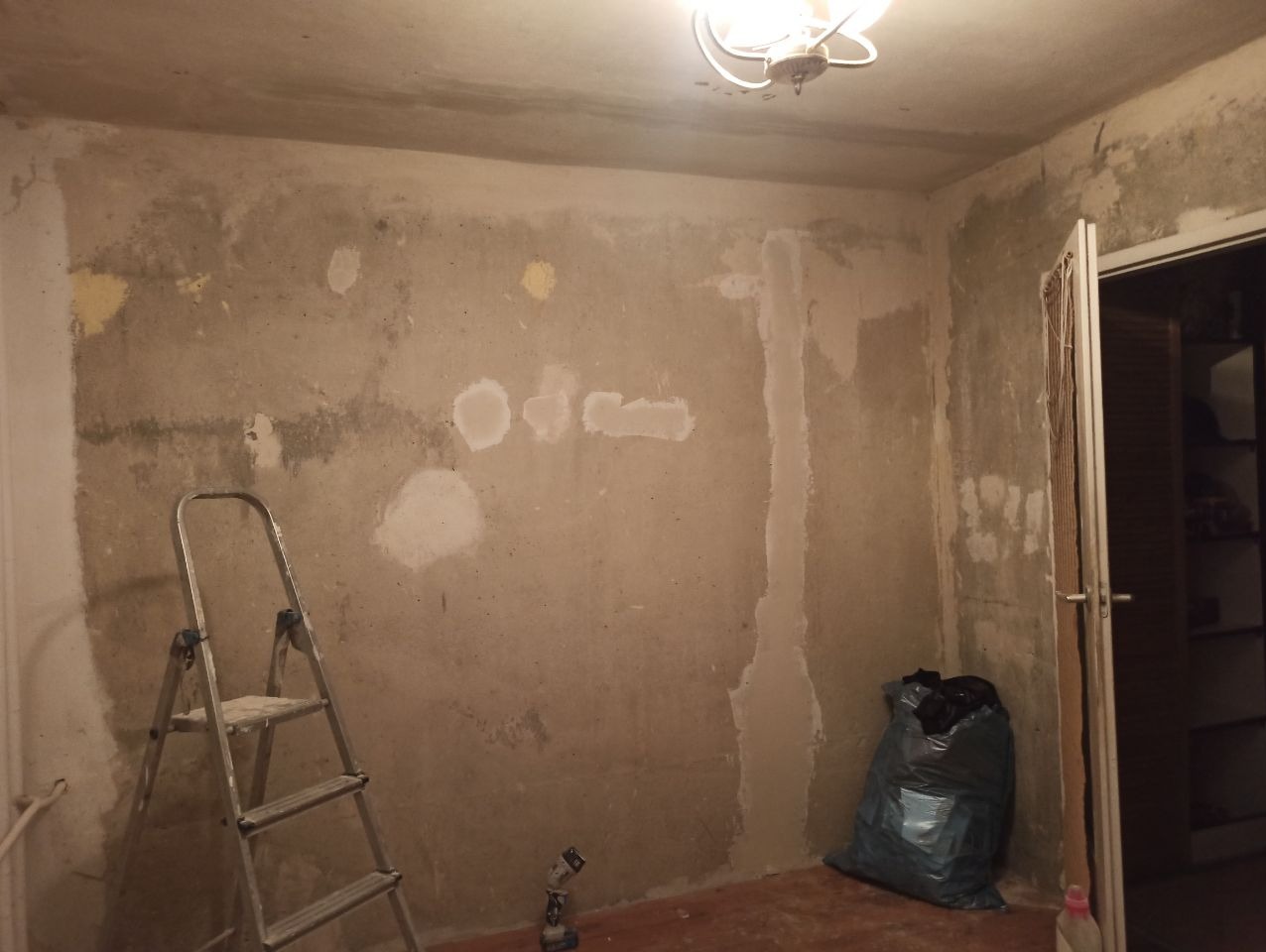 Pokój w trakcie przygotowania do malowania: ściany z widocznymi łatami gipsowymi, drabina, narzędzia i worki na śmieci. Widoczne niedoskonałości i surowy stan.
