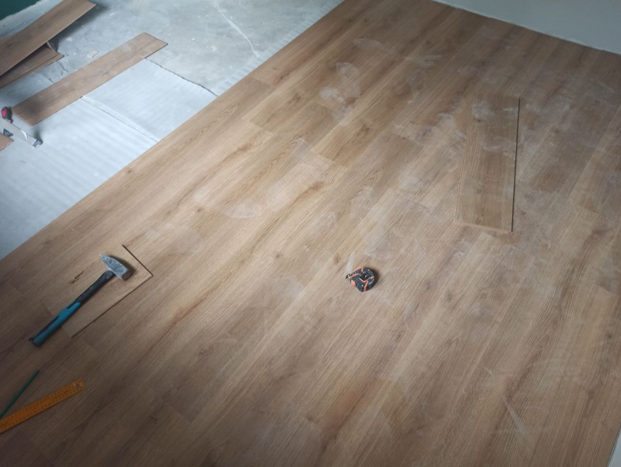 Układanie paneli podłogowych: widok z góry na częściowo ułożoną podłogę z paneli, z widocznym młotkiem, miarką i fragmentami nieułożonych paneli w tle.