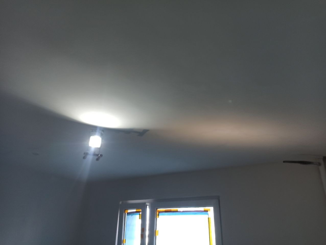 Biały sufit po malowaniu z widoczną lampą halogenową i oknem w tle. Świeżo pomalowane ściany w jasnym odcieniu. Widoczne ślady po taśmie malarskiej na oknach.