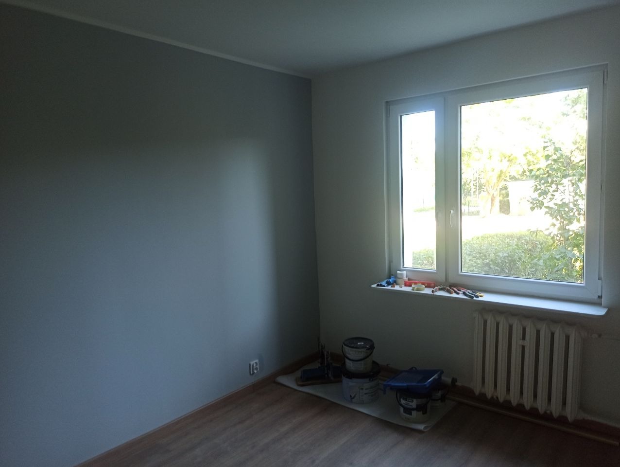 Pomieszczenie po malowaniu ścian na szaro, widoczne narzędzia malarskie przy oknie i grzejniku. Podłoga drewniana, okno z widokiem na zieleń.