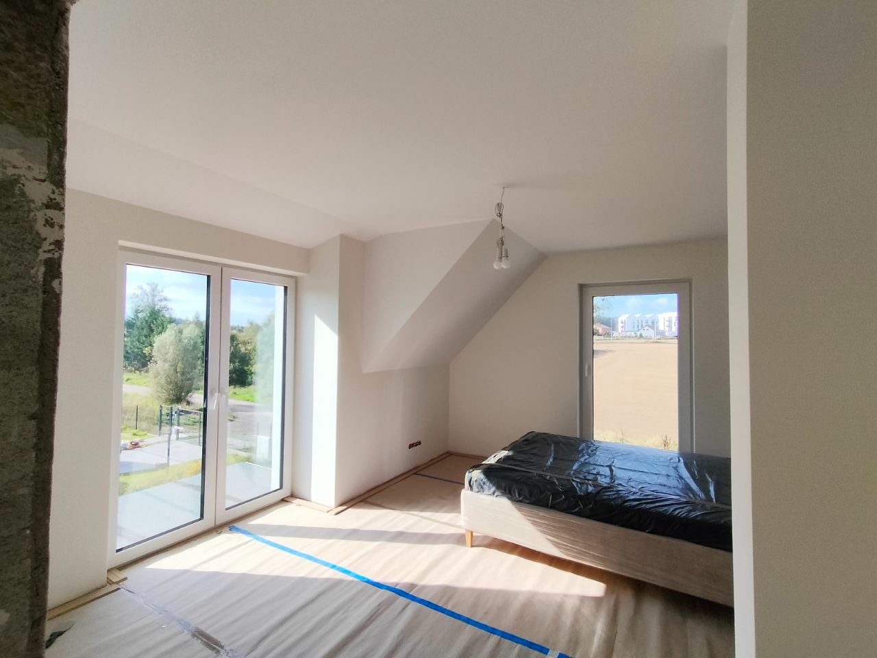 Jasne poddasze po malowaniu ścian na biało, z oknami i zabezpieczonym łóżkiem folią. Podłoga zakryta folią malarską z niebieską taśmą. Widok na zewnątrz przez okna.