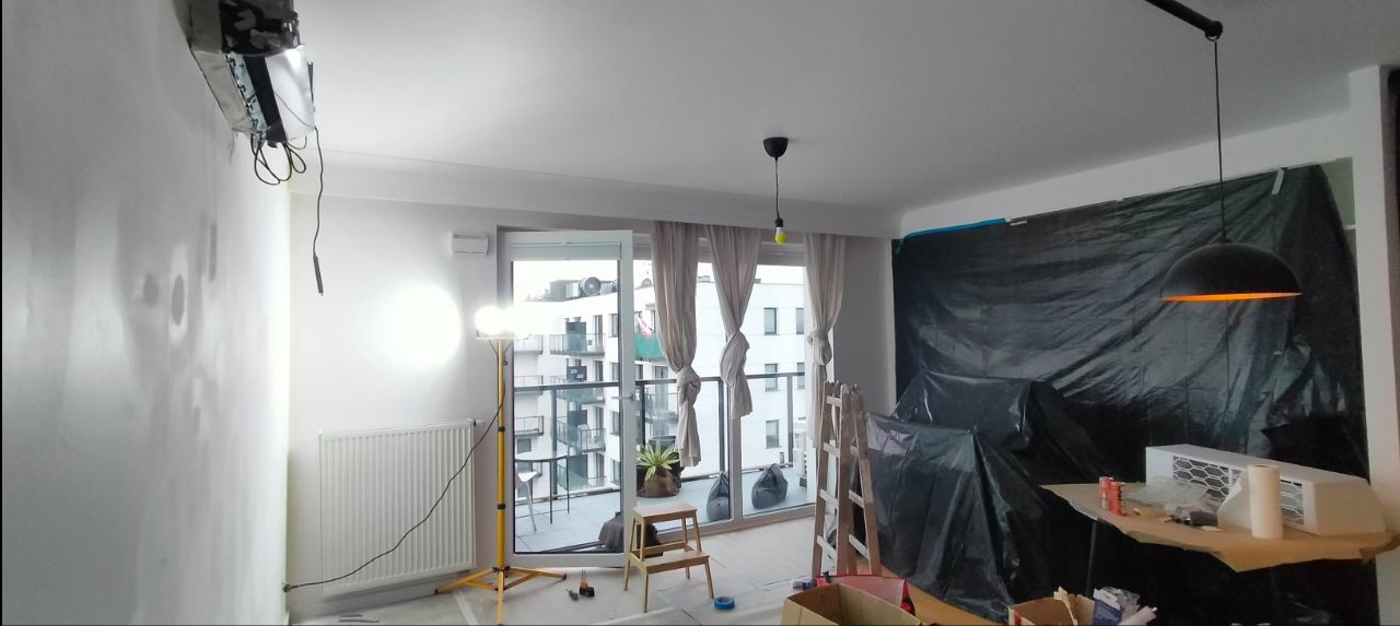 Remont mieszkania w trakcie: zabezpieczone folią meble, przygotowana ściana do malowania, widoczny grzejnik i lampa na statywie. Prace wykończeniowe w jasnym wnętrzu.