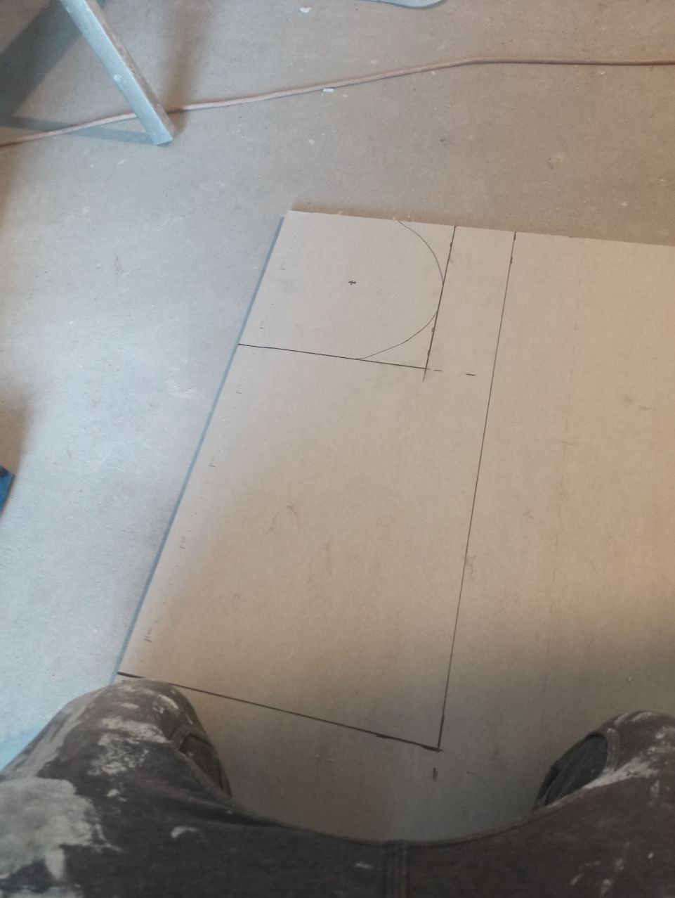 Płyta gipsowo-kartonowa z odrysowanym okręgiem i liniami cięcia, leżąca na podłodze, widok z góry, spodnie robocze w dolnej części kadru.