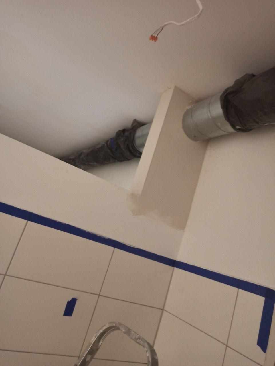 Kanciasty kanał wentylacyjny przymocowany do sufitu i ściany, zabezpieczone niebieską taśmą malarską, widoczne płytki i fragment drabiny.