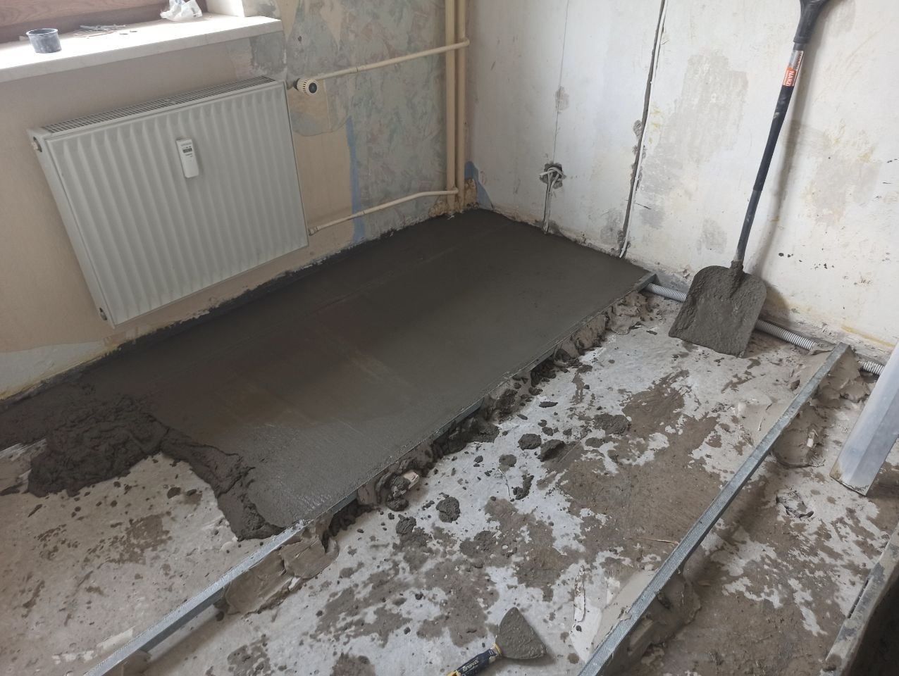 Wyrównywanie podłoża wylewką samopoziomującą w trakcie remontu. Widoczna łopata i listwy prowadzące. Ściany w stanie surowym, grzejnik na ścianie.