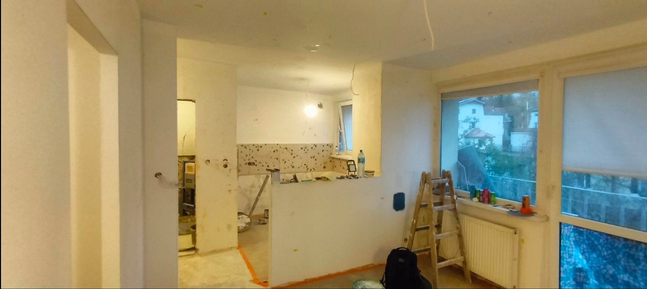 Wnętrze w trakcie remontu: widoczne ściany przygotowane do malowania, częściowo położone płytki w kuchni, drabina oparta o okno. Etap prac wykończeniowych.