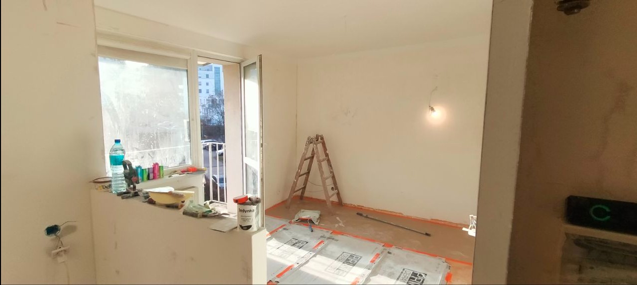 Pokój w trakcie malowania: drabina, farba Jedynka, zabezpieczona podłoga, widok na balkon z butelką wody. Przygotowania do malowania ścian w jasnym wnętrzu.