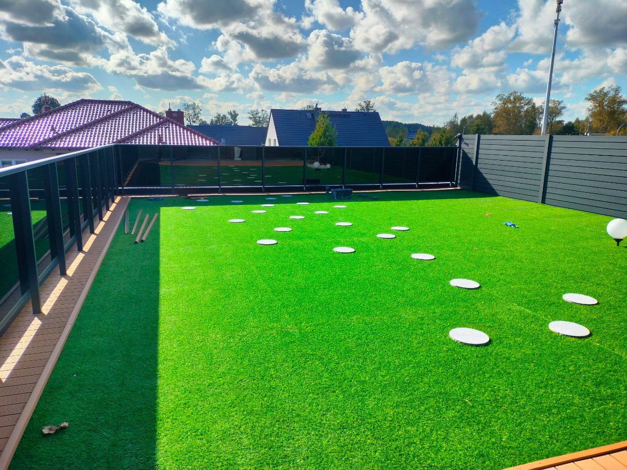 Nowoczesny ogród z zieloną sztuczną trawą, białymi okrągłymi płytami chodnikowymi i ciemnym ogrodzeniem panelowym. Widok na domy z czerwonymi dachami w tle.