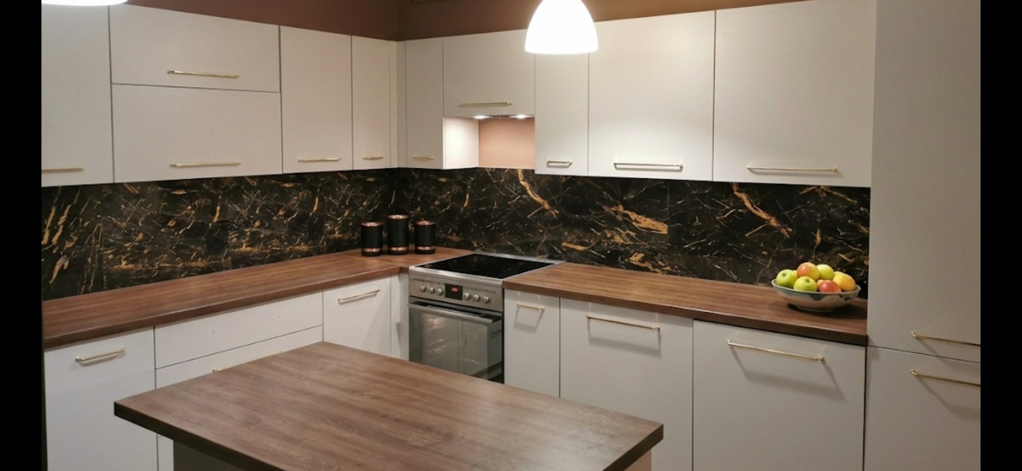 Nowoczesna kuchnia z białymi szafkami, drewnianym blatem i marmurowym panelem ściennym. Na blacie misa z owocami i zestaw pojemników. Oświetlenie sufitowe.