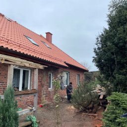 USŁUGI DEKARSKO-BUDOWLANE MARCIN CICHOWSKI - Czerwony dach z dachówkami ceramicznymi i oknami połaciowymi na ceglanym domu. Widoczny fragment drewnianej konstrukcji i osoba pracująca na zewnątrz.