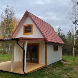 USŁUGI DEKARSKO-BUDOWLANE MARCIN CICHOWSKI - Niewielki, biały domek z czerwonym dachem z blachodachówki, drewnianą werandą i brązowymi rynnami na tle jesiennego lasu pod pochmurnym niebem.