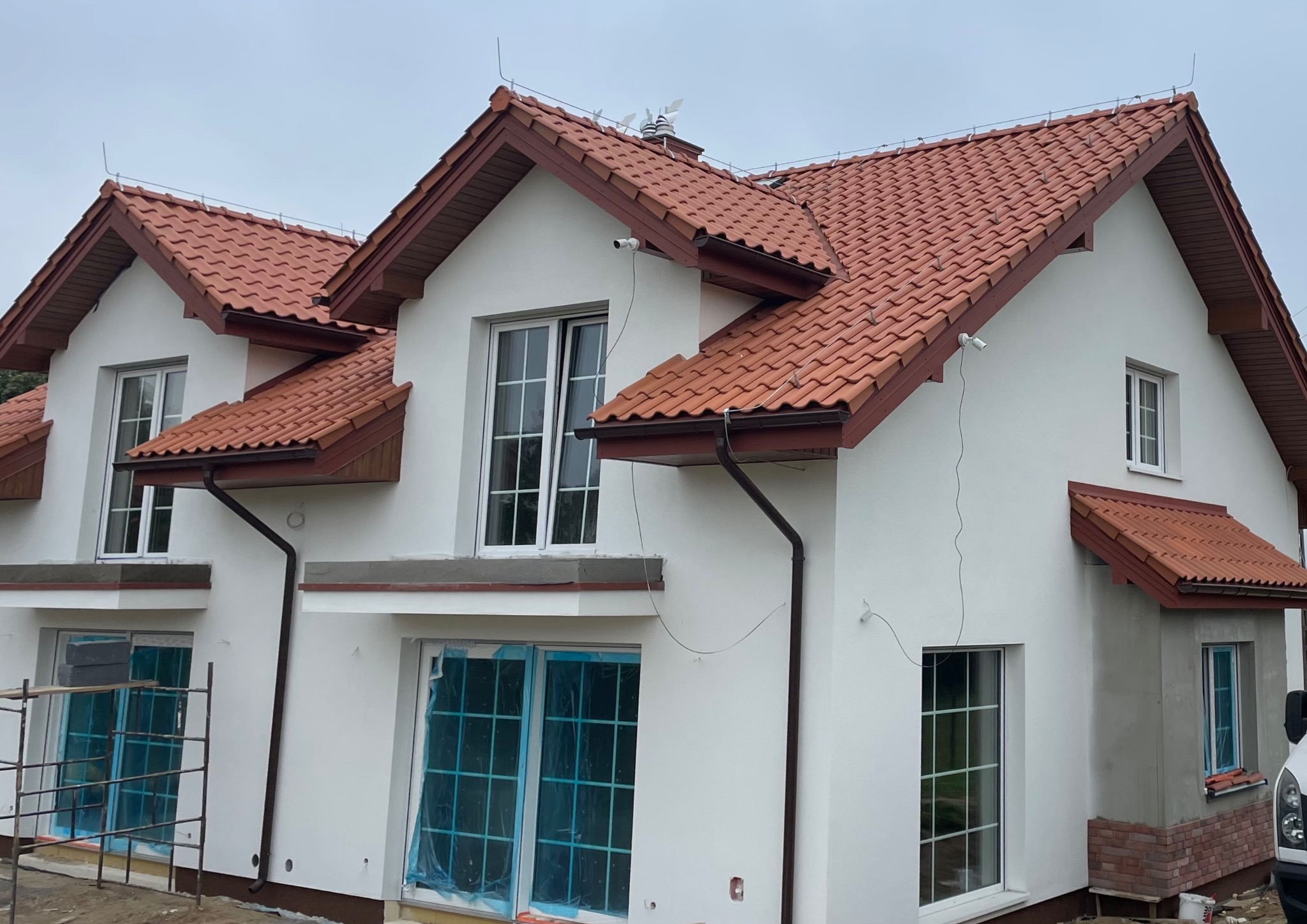 Nowoczesny dom z czerwoną dachówką, białymi ścianami i brązowymi rynnami. Widoczne okna zabezpieczone folią, elementy rusztowania i kamery monitoringu na elewacji.