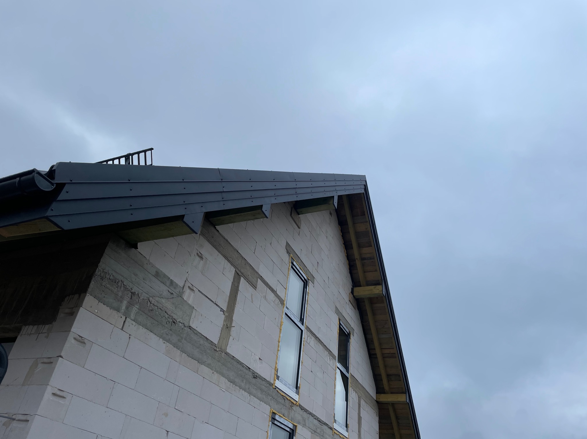 Surowy stan domu z czarnym dachem i rynnami pod pochmurnym niebem. Widoczne okna w trakcie montażu i elementy konstrukcyjne dachu.