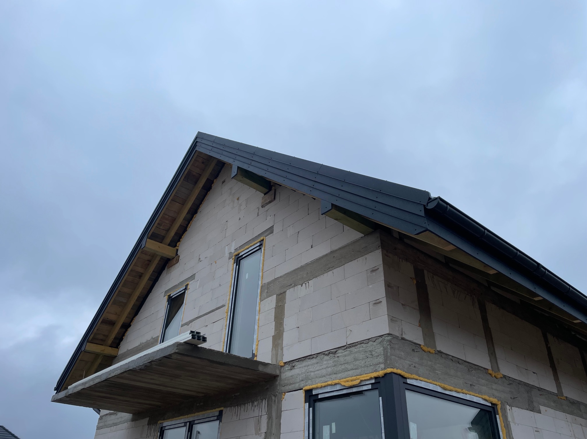 Niski kąt na dom w budowie z czarnym dachem i oknami. Widoczne białe bloczki, szare nadproża i żółta pianka montażowa. Pochmurne niebo w tle.