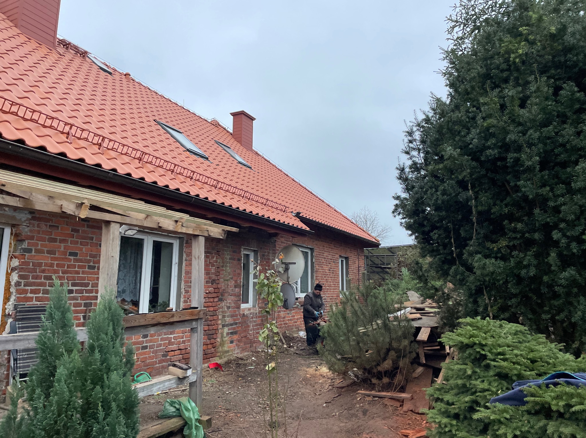 Czerwony dach z dachówkami ceramicznymi i oknami połaciowymi na ceglanym domu. Widoczny fragment drewnianej konstrukcji i osoba pracująca na zewnątrz.