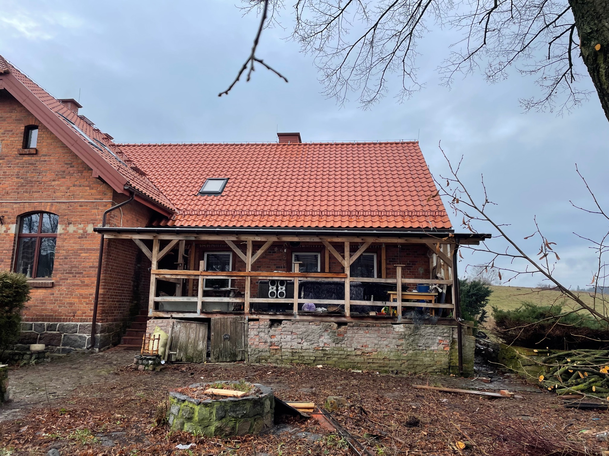 Ceglany dom z czerwonym dachem krytym dachówką, widoczne okna dachowe. Część domu z werandą w trakcie remontu, z drewnianą konstrukcją. Na pierwszym planie studnia.