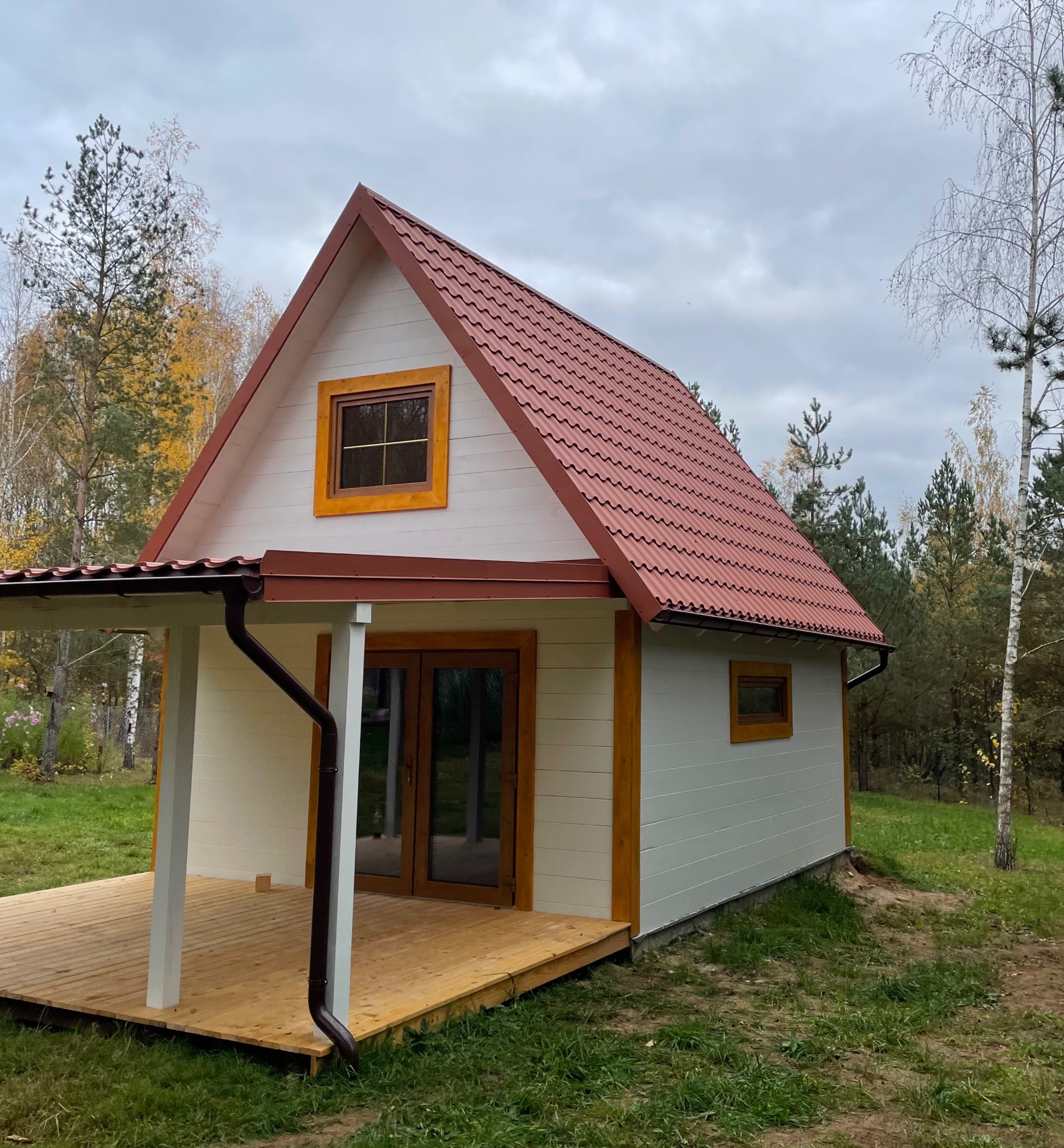 Niewielki, biały domek z czerwonym dachem z blachodachówki, drewnianą werandą i brązowymi rynnami na tle jesiennego lasu pod pochmurnym niebem.