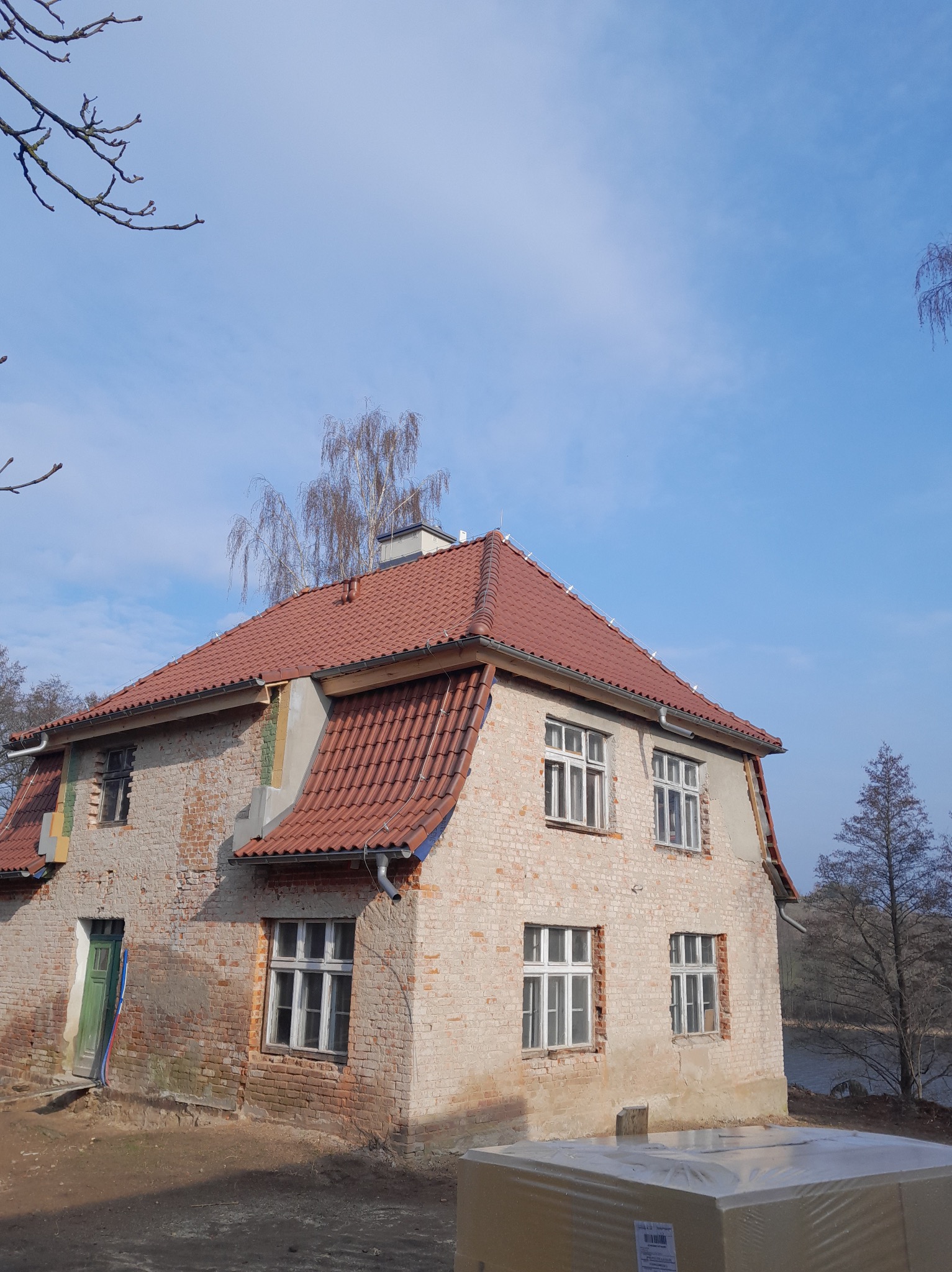 Dwukondygnacyjny budynek z czerwonym dachem z dachówki, częściowo otynkowany, z widoczną cegłą. Widoczne rynny i okna z białymi ramami. W tle drzewa i błękitne niebo.