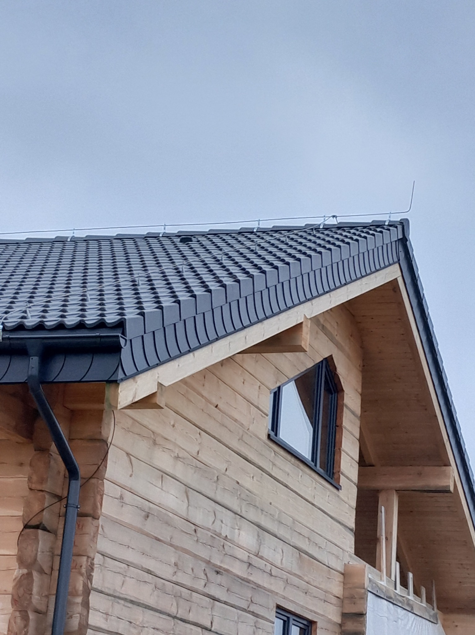 Fragment drewnianego domu z ciemnoszarym dachem z dachówki bitumicznej i rynną. Widoczne okno o nietypowym kształcie i system odgromowy na dachu.