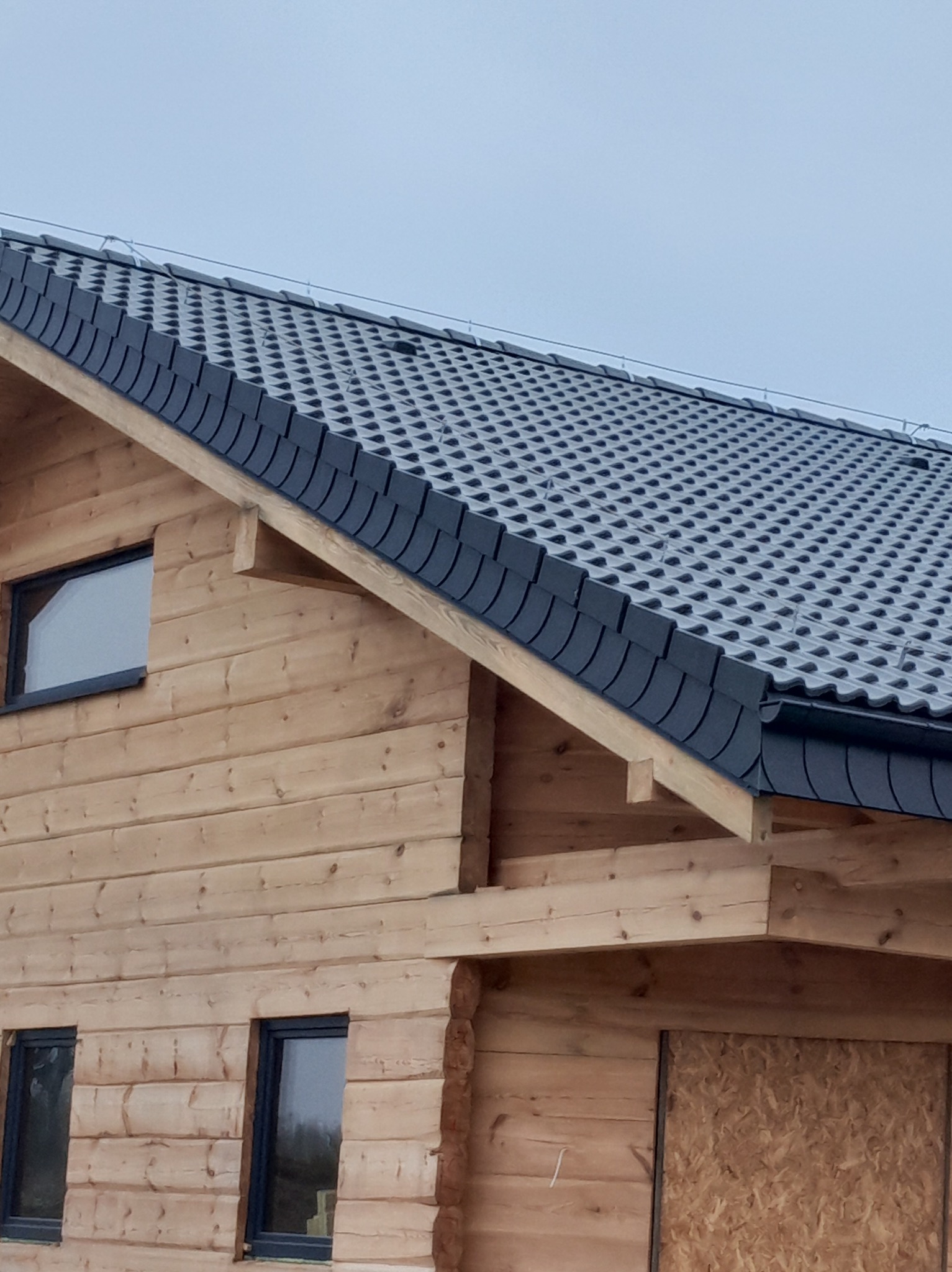 Dom z bali z nowym, ciemnoszarym dachem z dachówki karpiówki. Widoczne okna z czarnymi ramami i fragment ściany z płyt OSB.