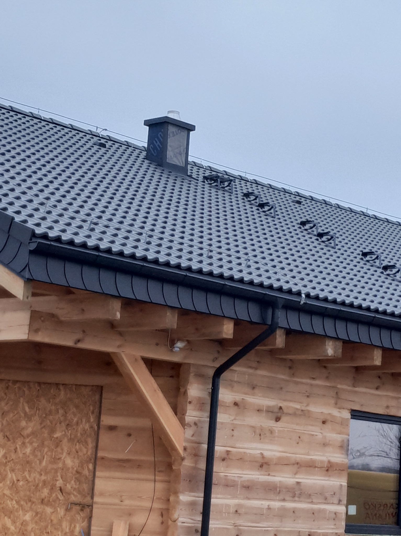 Szary dach z blachodachówki na drewnianym domu z kominem i rynną. Widoczne zabezpieczenia przeciwśniegowe.