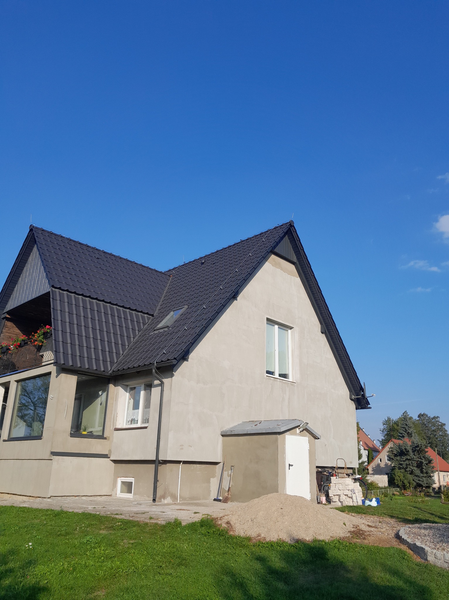 Dom jednorodzinny z ciemną dachówką, widok z boku na tle błękitnego nieba. Na balkonie widoczne kwiaty. Ściany budynku w trakcie wykończenia.