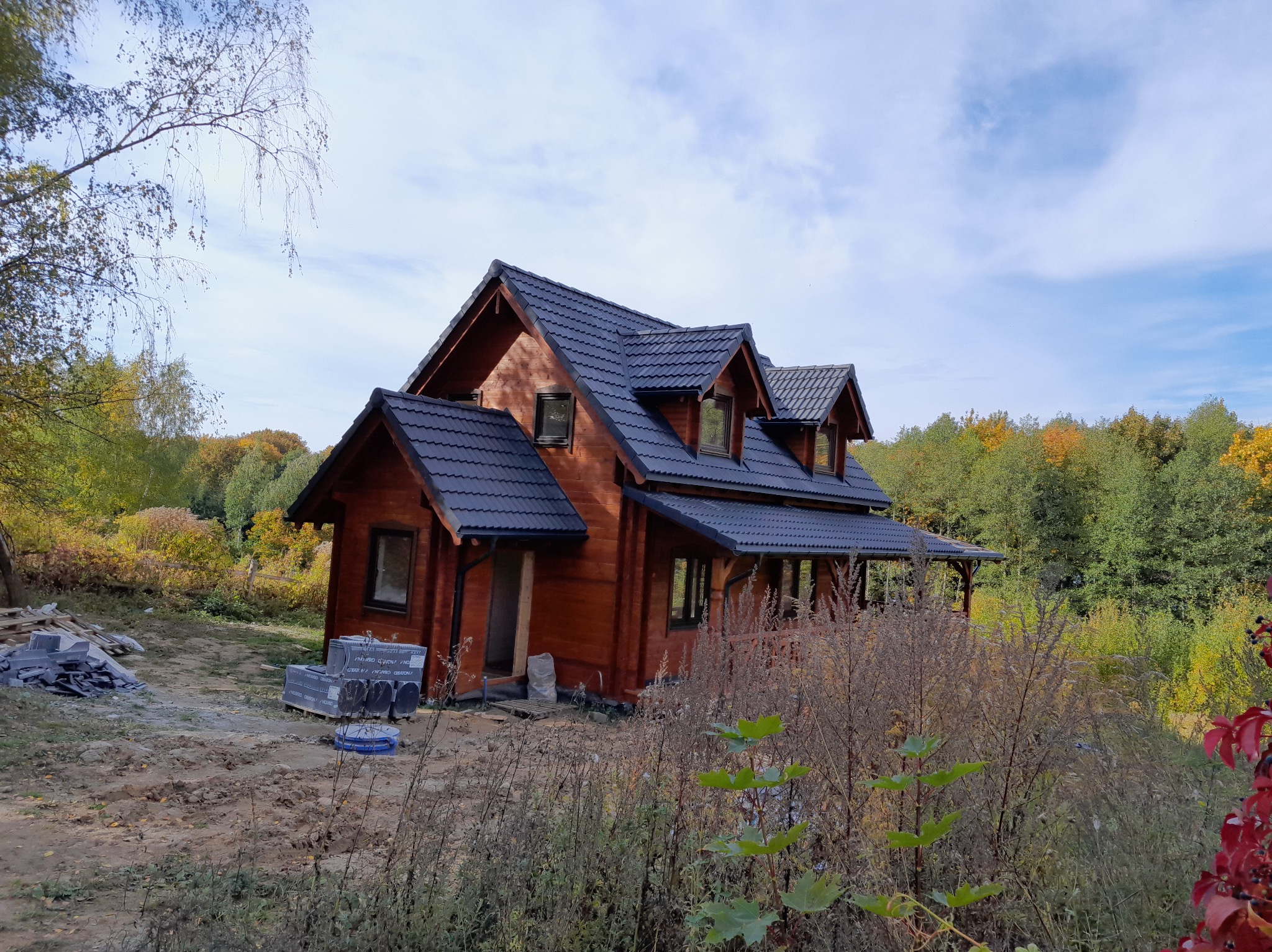 Drewniany dom z ciemnym dachem wielospadowym w Mrągowie, otoczony zielenią. Widoczne palety z dachówkami, co sugeruje prace dekarskie w toku.
