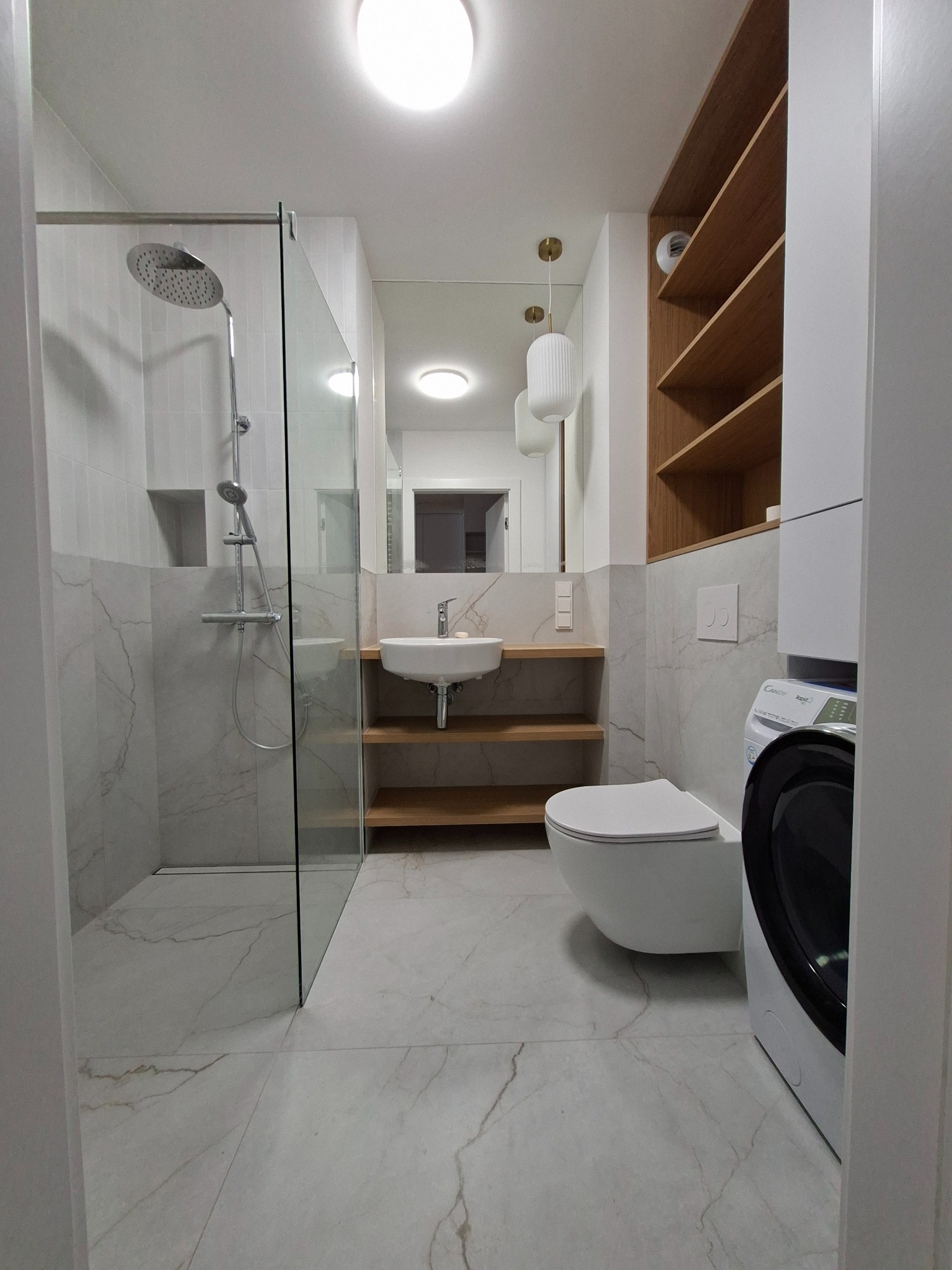 Jasna łazienka z kabiną prysznicową typu walk-in, umywalką na drewnianych półkach i pralką. Marmurowe płytki na podłodze i ścianach.