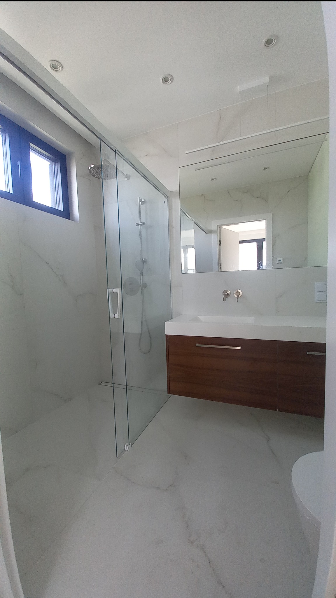 Nowoczesna łazienka z kabiną prysznicową typu walk-in, marmurowymi płytkami i drewnianą szafką pod umywalką. Minimalistyczny design z oświetleniem punktowym.