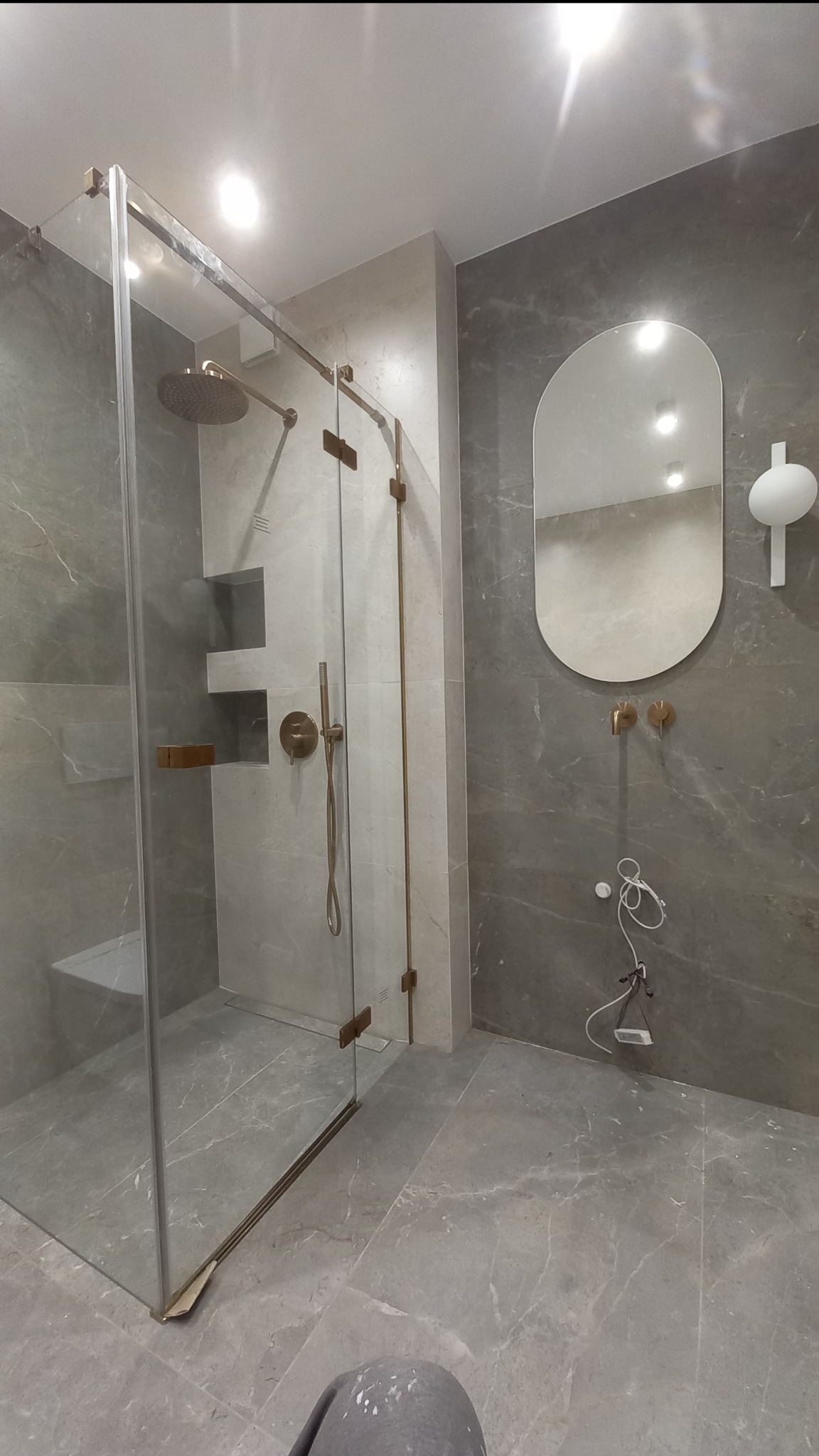 Nowoczesna łazienka z kabiną prysznicową typu walk-in, szare płytki imitujące marmur, owalne lustro z oświetleniem i złotymi akcentami armatury.