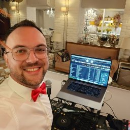 Karol Giżewski Music & Events - Uśmiechnięty DJ w muszce, z laptopem i sprzętem DJ-skim na tle eleganckiego wnętrza z dekoracjami. Widoczny mikrofon i elementy nagłośnienia.