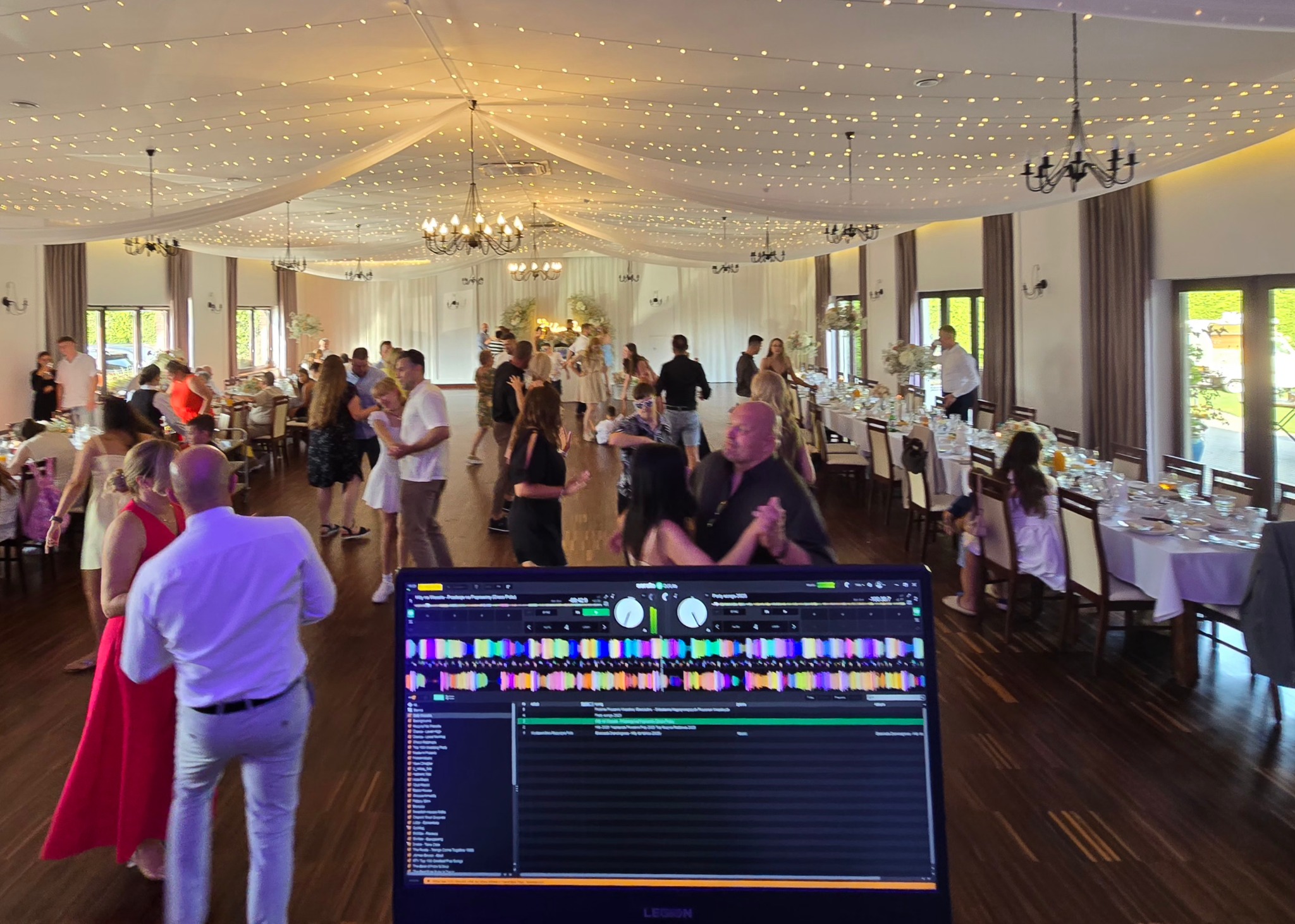Sala balowa z bawiącymi się gośćmi, oświetlona girlandami. W pierwszym planie widoczny ekran laptopa z oprogramowaniem DJ-skim, sugerujący oprawę muzyczną imprezy.