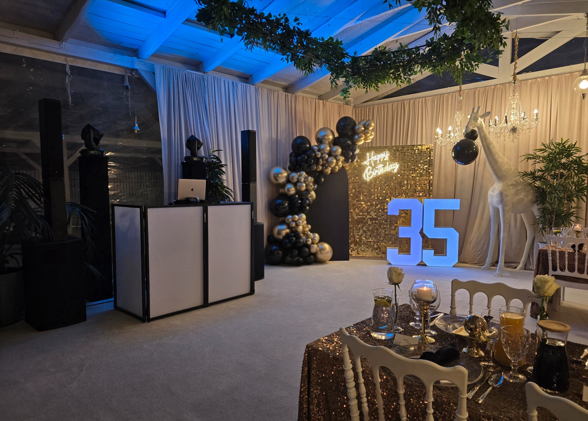 Dekoracja sali na 35 urodziny: złote cekiny, balony, podświetlane cyfry, żyrafa i DJ-ka z nagłośnieniem. Elegancki wystrój z elementami glamour i nowoczesnym oświetleniem.