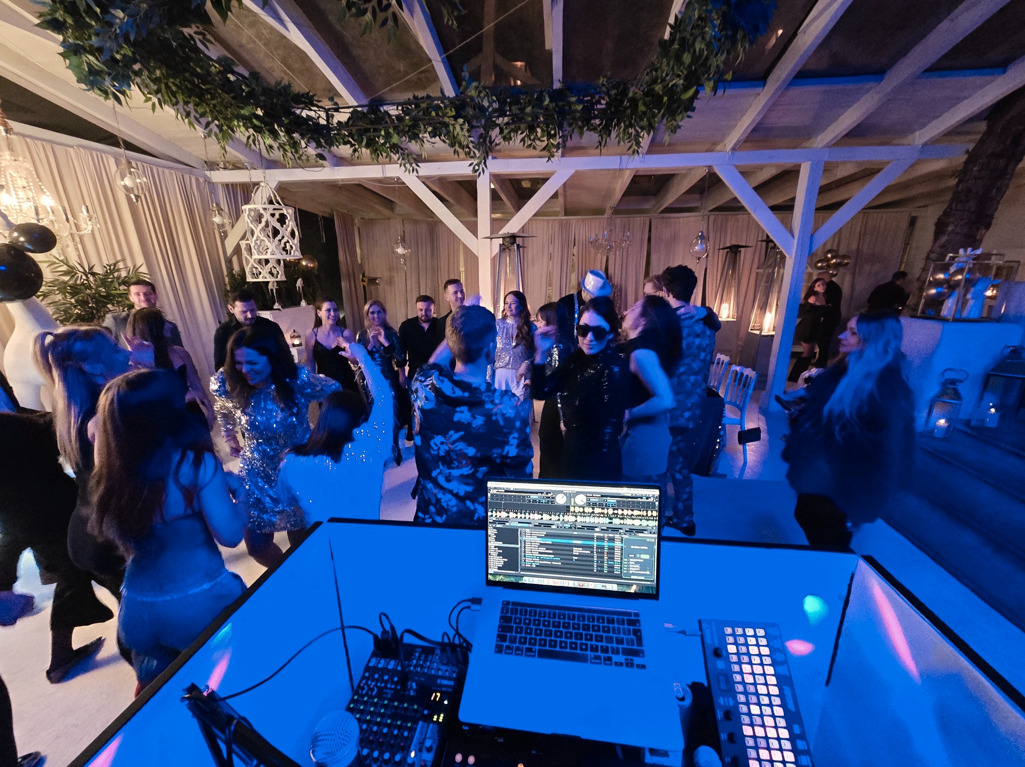 DJ grający muzykę na imprezie pod zadaszeniem, widoczny sprzęt DJ-ski i tańczący ludzie w tle, oświetlenie w odcieniach niebieskiego.