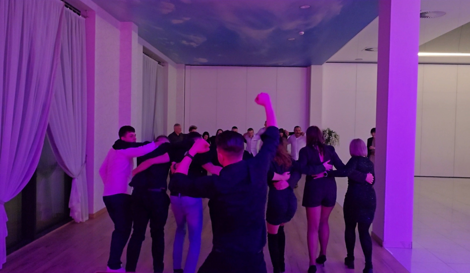 Rozbawieni goście weselni tańczą w kręgu w sali z fioletowym oświetleniem i sufitem imitującym niebo. Widoczni ludzie w różnym wieku, ubrani w eleganckie stroje.